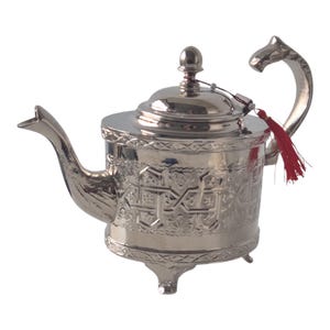 Op de afbeelding: Een zilverkleurige theepot met ingewikkelde reliëfpatronen en een decoratieve handgreep in de vorm van een dierenkop. De theepot heeft een tuit, een deksel met een knop en een kleine rode kwast. Hij staat op drie kleine voetjes.