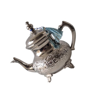 Op de afbeelding: Een zilveren theepot met ingewikkelde gegraveerde patronen en een gebogen handvat. De theepot heeft een deksel met een kleine knop en een blauwe kwast. De theepot is gemaakt van metaal en heeft drie kleine voetjes.
