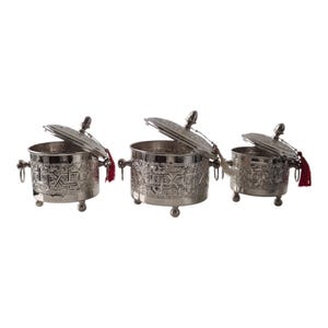 Op de afbeelding: Een set van drie zilverkleurige metalen bakjes met deksels, elk voorzien van ingewikkelde reliëf geometrische patronen, kleine bolvormige voetjes, ringvormige handgrepen aan de zijkant en een decoratief rood kwastje aan het deksel.