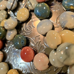 May include: A selection of colourful, glossy ceramic beads in shades of red, yellow, blue, and white. The beads are round and seem to be strung together. The background is a silver, decorative surface.