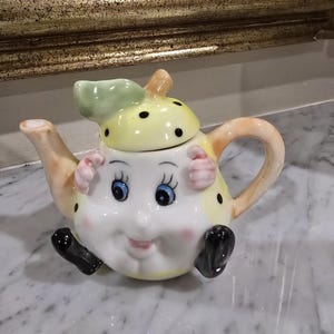 May include: A ceramic teapot shaped like a character with a smiling face, blue eyes, and pink cheeks. The teapot features a yellow lid with black polka dots and a green leaf handle, a curved spout, a peach-coloured handle, and black feet.
