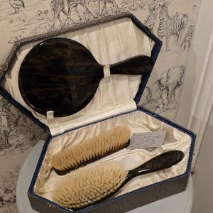May include: A vanity set in a hexagonal case featuring a dark brown tortoiseshell-patterned hand mirror and two hairbrushes with light-coloured bristles. The interior is lined with cream-coloured fabric, and a small handwritten note is tucked inside.