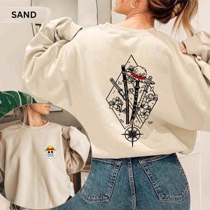 May include: Sand-colored sweatshirt with a graphic on the back featuring a straw hat, swords, and floral elements within a diamond shape. The front has a small skull and crossbones design. The word "SAND" is at the top.