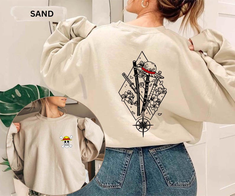 May include: Sand-colored sweatshirt with a graphic on the back featuring a straw hat, swords, and floral elements within a diamond shape. The front has a small skull and crossbones design. The word "SAND" is at the top.