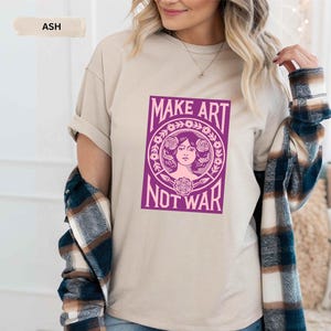 以下が含まれることがあります： 「MAKE ART NOT WAR」と書かれた紫色のグラフィックが特徴の、アッシュカラーのTシャツ。 グラフィックは、花の要素に囲まれた女性の顔が特徴です。 ゆったりとしたフィット感のシャツです。
