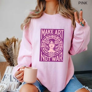 以下が含まれることがあります： ピンク色のスウェットシャツ。紫色のグラフィックで「MAKE ART NOT WAR」と書かれています。グラフィックは、花柄に囲まれた女性の顔が特徴です。スウェットシャツは無地の明るいピンク色です。
