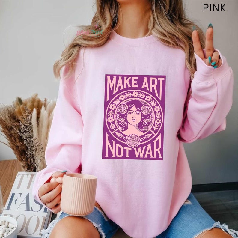 以下が含まれることがあります： ピンク色のスウェットシャツ。紫色のグラフィックで「MAKE ART NOT WAR」と書かれています。グラフィックは、花柄に囲まれた女性の顔が特徴です。スウェットシャツは無地の明るいピンク色です。