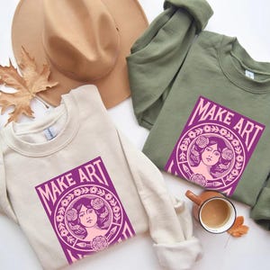 以下が含まれることがあります： クリーム色とオリーブグリーンの2枚のスウェットシャツ。それぞれに「MAKE ART」と書かれた紫色のグラフィックがプリントされています。グラフィックは、花に囲まれた女性の顔が特徴です。茶色の帽子、コーヒーカップ、秋の葉も画像にあります。