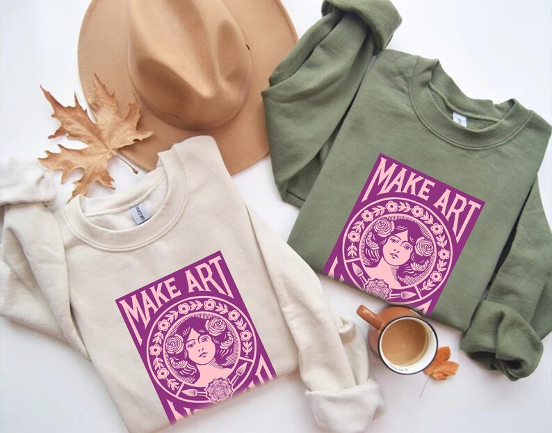 以下が含まれることがあります： クリーム色とオリーブグリーンの2枚のスウェットシャツ。それぞれに「MAKE ART」と書かれた紫色のグラフィックがプリントされています。グラフィックは、花に囲まれた女性の顔が特徴です。茶色の帽子、コーヒーカップ、秋の葉も画像にあります。