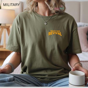 Op de afbeelding: Olijfgroen T-shirt met de tekst "TREATY OAK REVIVAL" in oranje. Het shirt heeft een relaxte pasvorm en korte mouwen. Het model draagt het shirt met een blauwe jeans.