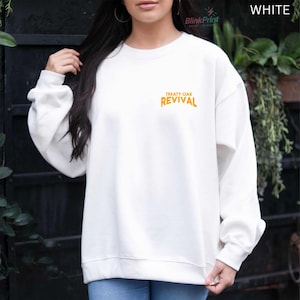 Op de afbeelding: Witte sweatshirt met de tekst "TREATY OAK REVIVAL" in oranje letters. De sweatshirt heeft een ronde hals en lijkt gemaakt van een zacht materiaal. Het woord "WHITE" is rechtsboven zichtbaar.