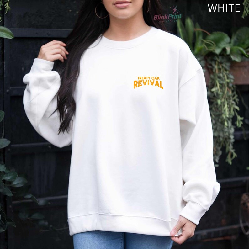 Op de afbeelding: Witte sweatshirt met de tekst "TREATY OAK REVIVAL" in oranje letters. De sweatshirt heeft een ronde hals en lijkt gemaakt van een zacht materiaal. Het woord "WHITE" is rechtsboven zichtbaar.