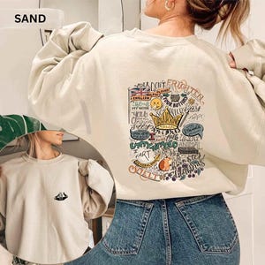 May include: Sand-colored sweatshirt with a graphic design on the back featuring a crown, a guinea pig, and various phrases. The sweatshirt has long sleeves and a crew neck. The word "SAND" is printed on the top left.