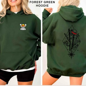 May include: Forest green hoodie with a front pocket and a hood. The front features a small skull and crossbones logo with a yellow hat. The back displays a black graphic of a hat, swords, and flowers within a diamond shape.