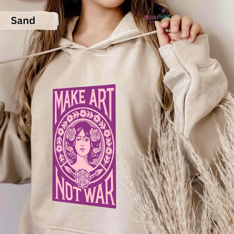 以下が含まれることがあります： 「MAKE ART NOT WAR」と書かれた紫色のグラフィックが特徴のサンドカラーのパーカー。グラフィックは、花の要素に囲まれた女性の顔が特徴です。パーカーにはドローストリングと長袖が付いています。