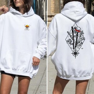May include: White hoodie with a front pocket and a hood. The front features a small, colorful skull and crossbones logo with a yellow hat. The back displays a black and white graphic of a hat, swords, and flowers within a diamond shape.