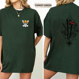 May include: Forest green t-shirt with a small skull and crossbones graphic on the front. The back of the shirt features a black graphic design with a hat, swords, and a compass rose. The shirt is a relaxed fit.