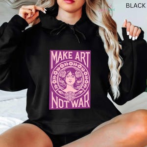 以下が含まれることがあります： 「MAKE ART NOT WAR」の文字と女性の肖像画が描かれた紫色のグラフィックが特徴の黒いパーカー。 パーカーには、ドローストリング付きのフードと長袖が付いています。