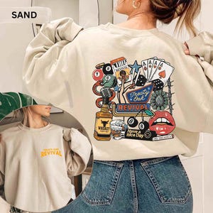 Op de afbeelding: Zandkleurige sweatshirt met een afbeelding op de achterkant met een fles whisky, speelkaarten, dobbelstenen en de woorden "Treaty Oak Revival". De voorkant van de sweatshirt heeft de woorden "Treaty Oak Revival" in oranje.