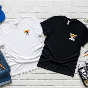 May include: Two t-shirts, one white and one black, each with a small graphic of a skull and crossbones wearing a straw hat. A blue baseball cap, denim jeans, and black and white sneakers are also visible.
