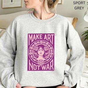 以下が含まれることがあります： 「MAKE ART NOT WAR」と書かれた紫色のグラフィックが特徴の、ライトグレーのスウェットシャツ。 グラフィックは、花柄とアートツールに囲まれた女性の顔が特徴です。 スウェットシャツはカジュアルで快適なスタイルです。