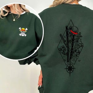 May include: Dark green sweatshirt with a white-trimmed neckline. The front features a small embroidered pirate skull and crossbones. The back displays a black graphic of a hat, swords, flowers, and a compass rose within a diamond.