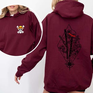 May include: Burgundy hoodie with a small skull and crossbones logo on the front and a large graphic on the back. The back graphic features a straw hat, swords, flowers, and a compass design. The hoodie has a hood and long sleeves.