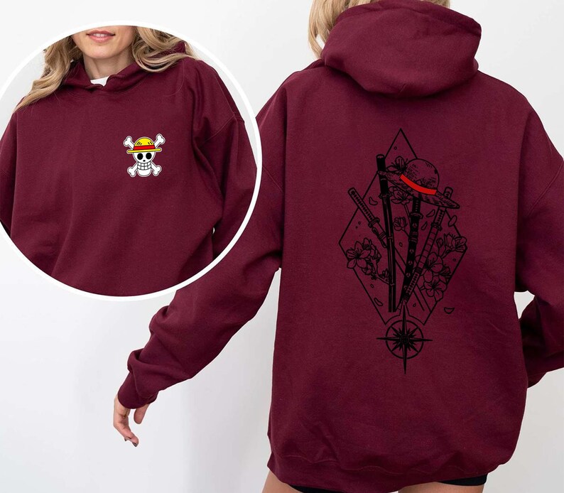 May include: Burgundy hoodie with a small skull and crossbones logo on the front and a large graphic on the back. The back graphic features a straw hat, swords, flowers, and a compass design. The hoodie has a hood and long sleeves.