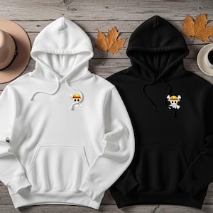 May include: Two hooded sweatshirts, one white and one black, with a front pocket and a small graphic of a skull and crossbones wearing a straw hat. The sweatshirts are on a wooden surface with autumn leaves and a hat.