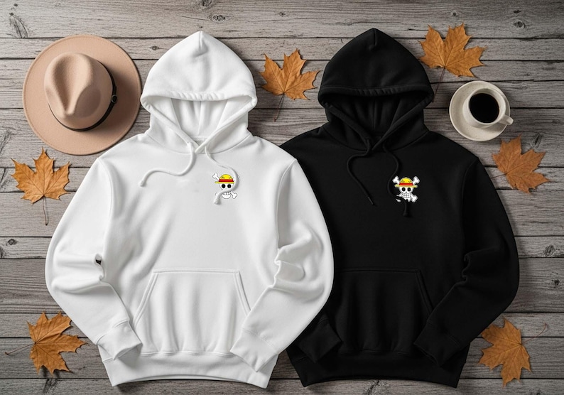May include: Two hooded sweatshirts, one white and one black, with a front pocket and a small graphic of a skull and crossbones wearing a straw hat. The sweatshirts are on a wooden surface with autumn leaves and a hat.