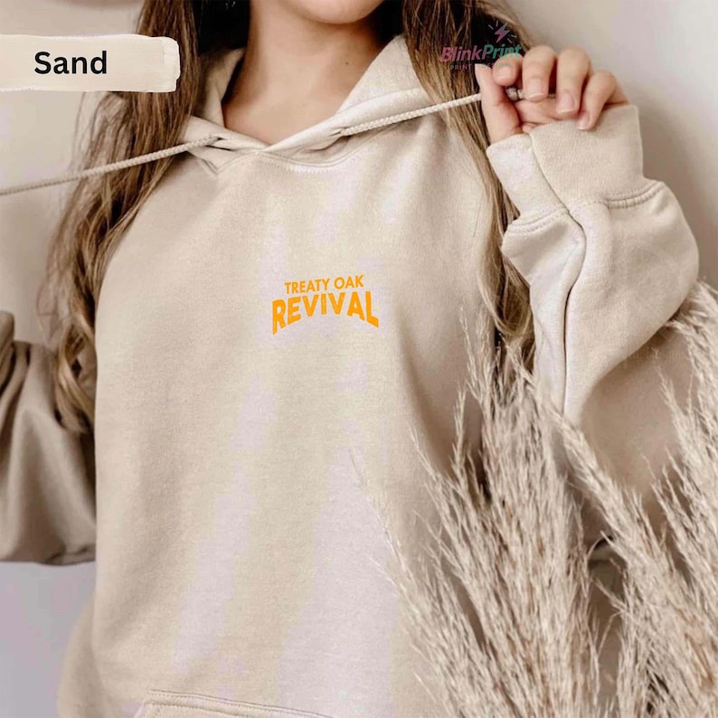 Op de afbeelding: Een zandkleurige hoodie met de oranje tekst "TREATY OAK REVIVAL". De hoodie heeft een trekkoord en een voorzak. De lange mouwen en het zachte materiaal suggereren een casual stijl. Het ontwerp is eenvoudig en comfortabel.