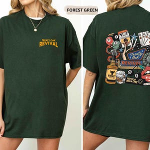 Op de afbeelding: Bosgroen t-shirt met de tekst "Treaty Oak Revival" op de voorkant en een afbeelding op de achterkant. De afbeelding op de achterkant bevat een whiskyfles, speelkaarten, een microfoon en de woorden "Treaty Oak Revival" en "Have a nice day."