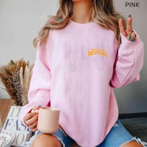 Op de afbeelding: Een lichtroze sweatshirt met de oranje tekst "TREATY OAK REVIVAL". De crewneck sweatshirt is effen van kleur. De afbeelding toont een persoon die de sweatshirt draagt.