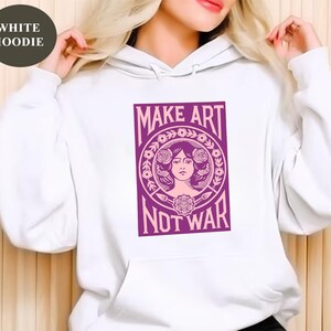 以下が含まれることがあります： 「MAKE ART NOT WAR」と書かれた紫色のグラフィックが特徴の白いパーカー。グラフィックは、花の要素に囲まれた女性の顔が特徴です。パーカーには、ドローストリングフードとフロントポケットが付いています。
