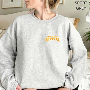 Op de afbeelding: Een lichtgrijs sweatshirt met de oranje tekst "TREATY OAK REVIVAL". De sweatshirt heeft een ronde hals en lijkt gemaakt van een zacht materiaal. De woorden "SPORT GREY" zijn rechtsboven zichtbaar.