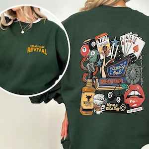 Op de afbeelding: Donkergroene sweatshirt met de tekst "Treaty Oak Revival" op de voor- en achterkant. De achterkant heeft een afbeelding met speelkaarten, dobbelstenen, een fles whisky en de tekst "Have a nice day."