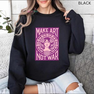 以下が含まれることがあります： 「MAKE ART NOT WAR」と書かれた紫色のグラフィックが特徴の黒いスウェットシャツ。 グラフィックは、花の要素に囲まれた女性の顔を特徴としています。 テキストは大胆で様式化されたフォントです。