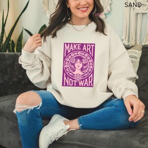 以下が含まれることがあります： 「MAKE ART NOT WAR」と書かれた紫色のグラフィックが特徴の、サンドカラーのスウェットシャツ。 グラフィックは、花の要素に囲まれた女性の顔が特徴です。 スウェットシャツは、ブルージーンズと白いスニーカーと組み合わせています。