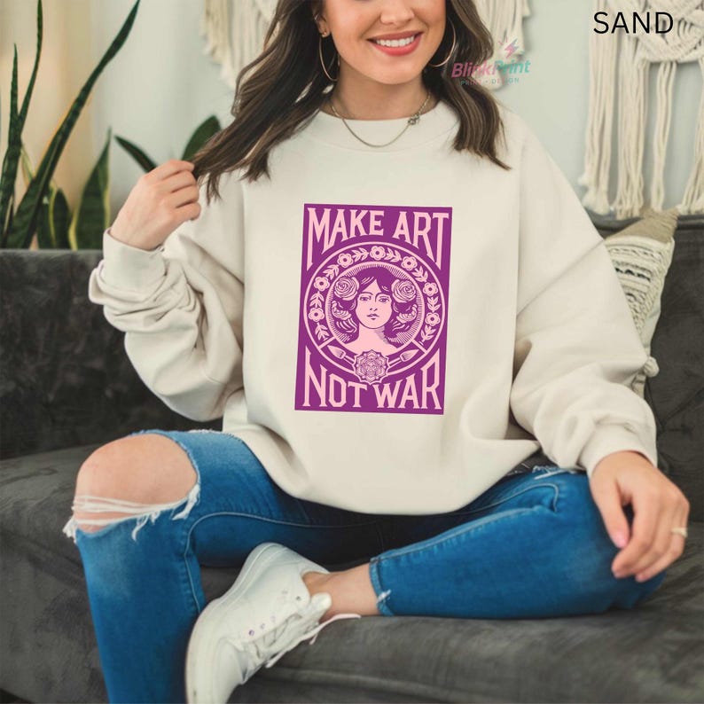 以下が含まれることがあります： 「MAKE ART NOT WAR」と書かれた紫色のグラフィックが特徴の、サンドカラーのスウェットシャツ。 グラフィックは、花の要素に囲まれた女性の顔が特徴です。 スウェットシャツは、ブルージーンズと白いスニーカーと組み合わせています。