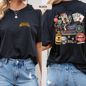 Op de afbeelding: Zwart T-shirt met korte mouwen met de tekst "Treaty Oak Revival" in goud op de voorkant. De achterkant van het shirt heeft een kleurrijke afbeelding met een fles whisky, speelkaarten en de woorden "Have a nice day."