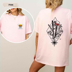 May include: Pink t-shirt featuring a small cartoon skull and crossbones with a straw hat on the front. The back of the shirt displays a black and white graphic of a straw hat, swords, flowers, and a compass within a diamond shape.