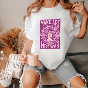 以下が含まれることがあります： 「MAKE ART NOT WAR」と書かれた紫色のグラフィックが特徴の白いTシャツ。グラフィックは、花の要素に囲まれた女性の顔が特徴です。このシャツはカジュアルなスタイルで、普段使いに適しています。