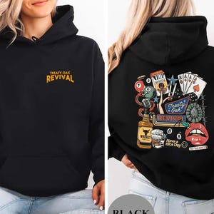 Op de afbeelding: Zwarte hoodie met een voorzak en een capuchon met trekkoord. De voorkant heeft de tekst "TREATY OAK REVIVAL" in oranje. De achterkant heeft een kleurrijke afbeelding met speelkaarten, een fles whisky en de tekst "Treaty Oak Revival."