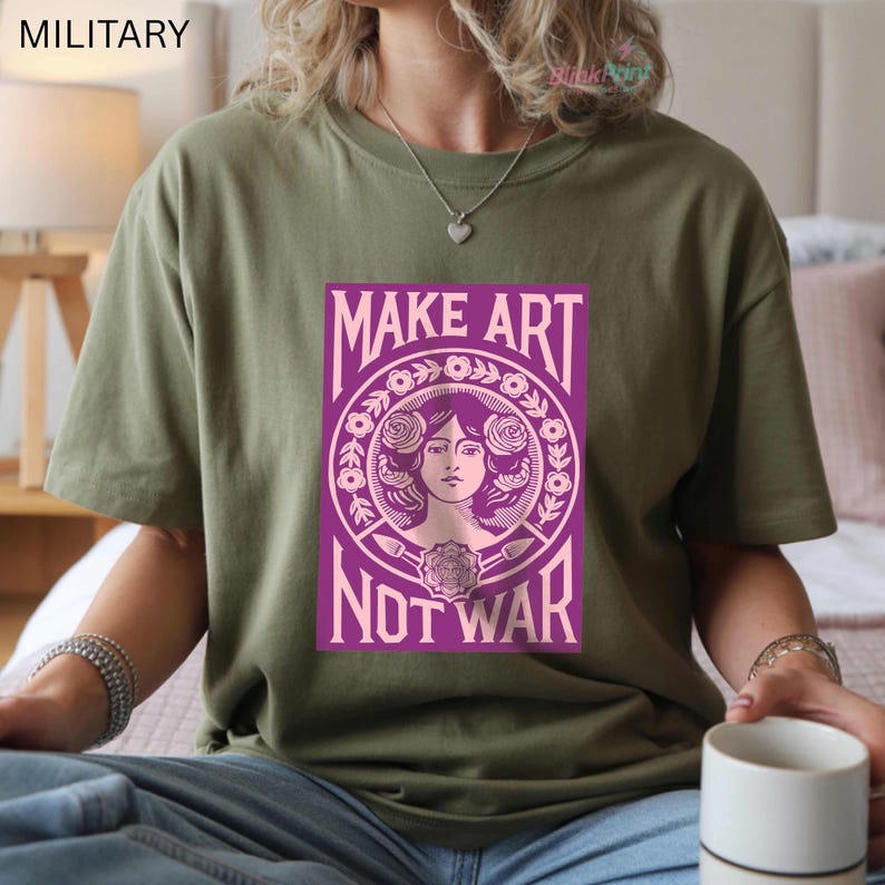 以下が含まれることがあります： オリーブグリーンのTシャツ。紫色のグラフィックで「MAKE ART NOT WAR」と書かれています。デザインは、花柄に囲まれた女性の肖像画です。カジュアルな普段着です。