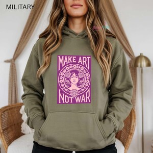 以下が含まれることがあります： オリーブグリーンのパーカー。紫色の文字で「MAKE ART NOT WAR」と書かれたグラフィックが前面にプリントされています。グラフィックは女性の顔と花の要素が特徴です。パーカーにはカンガルーポケットとドローストリングフードが付いています。