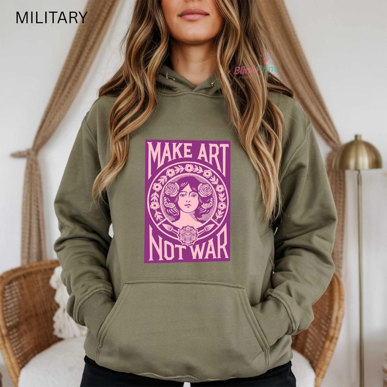 以下が含まれることがあります： オリーブグリーンのパーカー。紫色の文字で「MAKE ART NOT WAR」と書かれたグラフィックが前面にプリントされています。グラフィックは女性の顔と花の要素が特徴です。パーカーにはカンガルーポケットとドローストリングフードが付いています。