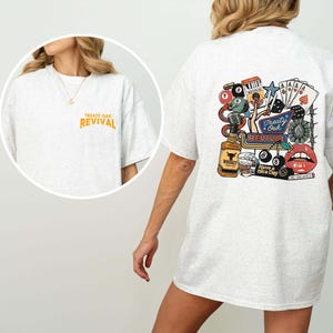 Op de afbeelding: Lichtgrijs t-shirt met de tekst "Treaty Oak Revival" in oranje op de voorkant en een kleurrijke afbeelding op de achterkant. De afbeelding bevat een whiskyfles, speelkaarten, dobbelstenen en een paar rode lippen.