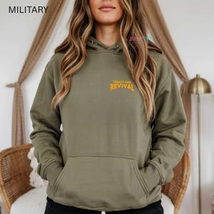 Op de afbeelding: Olijfgroene hoodie met een voorzak en een capuchon. De tekst "TREATY OAK REVIVAL" is in oranje op de borst gedrukt. Het woord "MILITARY" staat linksboven gedrukt.