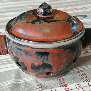 Peut inclure: Un pot à ail en céramique avec un couvercle. Le pot est émaillé dans des tons de rouge, d'orange et de noir, avec de petits trous pour la ventilation. Il a deux poignées et un petit bouton sur le dessus du couvercle.