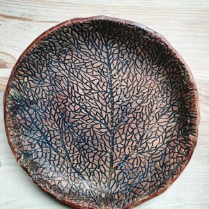 May include: A handmade, round ceramic plate with a leaf-like pattern. The plate has a dark brown rim and a textured surface with a network of black lines over a lighter brown base. The plate is approximately 25 cm in diameter.